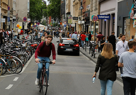 Foto der belebten Ehrenstraße an einem Samstagnachmittag. Radfahrende, zu Fuß Gehende teilen sich den Raum. Zudem ist ein Auto zu sehen, dass sich seinen Weg bahnt.