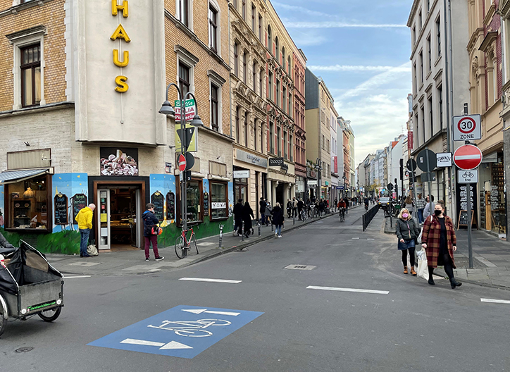 Foto von einer Straßenkreuzung eines typischen Kölner Wohnviertels mit Fahrradstraße und vielen Fußgängern, die unterwegs sind.