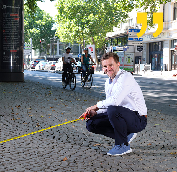Foto von Nico Rathmann, wie er in der Kölner Innenstadt lächelnd einen Fußweg mit einem Bandmaß ausmisst.